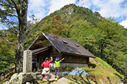 緑の山々を背景に、三角屋根の行者還小屋が建ち、2名の参加者が山を眺めている様子を後ろから撮影した写真