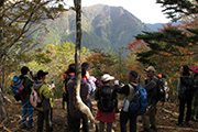 奥に大蛇ぐらの山並みが展望できる、周囲に立つ木々の葉が紅葉に色づいている展望所に大勢の登山者が集まっている写真