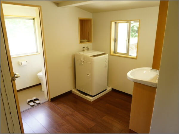 奥のドアにはトイレが設置され、手前の部屋には白い洗濯機と洗面台が設置された脱衣所内部の写真